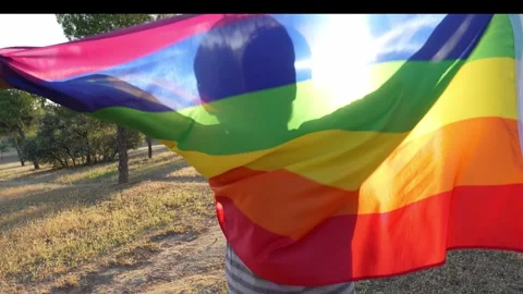 Non-binary guy waving rainbow flag with joy and spin backlighted in full sun 스톡 동영상 250468692