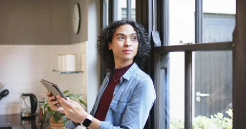 Non-binary individual tapping tablet with smartwatch at kitchen window gazing Stock Footage 335986048