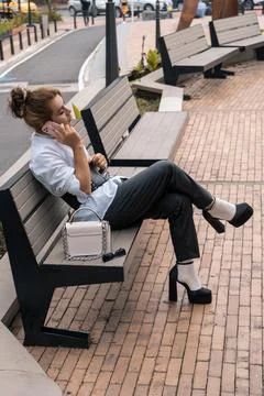Non-binary man talking using phone in her hands leaning on park bench in the Stock Photos