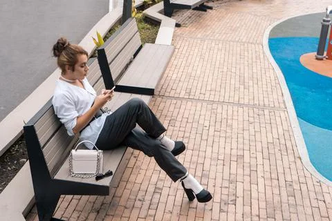 Non-binary man talking using phone in her hands leaning on park bench in the Stock Photos