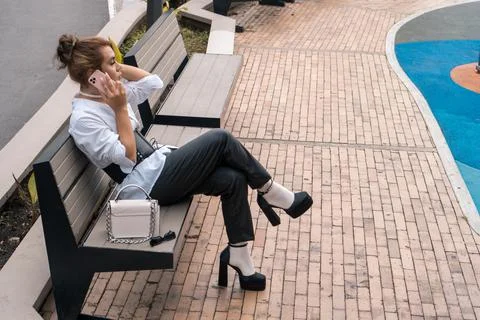 Non-binary man talking using phone in her hands leaning on park bench in the Stock Photos