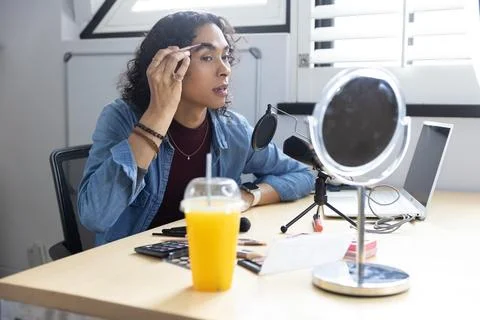 Non-binary mid-20s grooming eyebrows at home desk with mirror and mic using Stock Photos