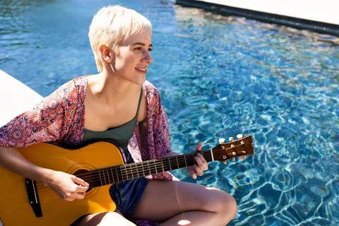 Non-binary musician sitting poolside in sun, playing acoustic guitar near clear Stock Photos