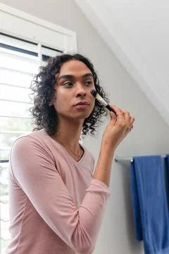 Non-binary person applying makeup brush to cheek in bathroom near white shutters Stock Photos