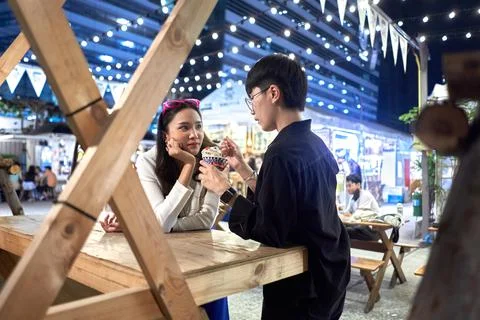 Non binary person feeding ice cream to her partner at urban night fair Stock Photos
