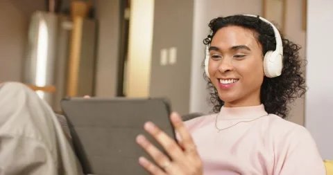 Non-binary person hearing audio through headphones and tapping tablet on sofa, Stock Footage 342492220