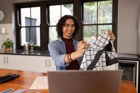 Non-binary person holding checkered top over laptop at kitchen counter with Stock Photos
