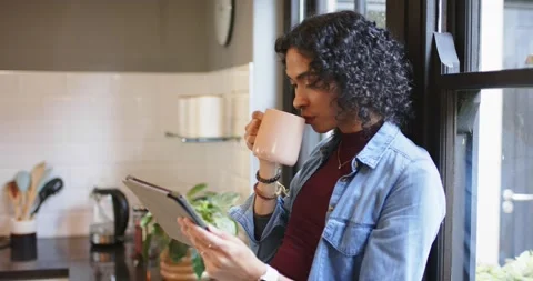 Non-binary person at kitchen window reading tablet sipping pink mug scrolling Stock Footage 337159592