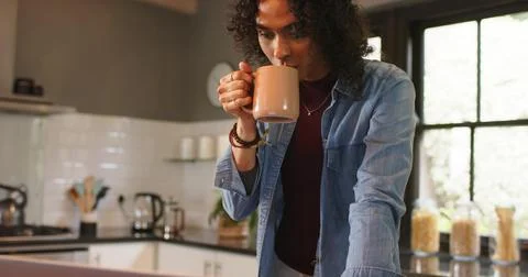 Non-binary person leaning at kitchen counter reviewing laptop reaching mug Stock Photos