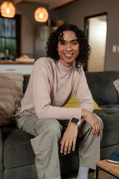 Non-binary person leaning on sofa smiling under woven pendant lamps by kitchen Stock Photos