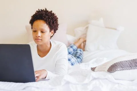 Non-binary person lying on bed in bedroom, working on laptop with white pillows, Stock Photos