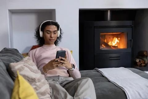 Non-binary person lying on gray sofa at home, wearing white headset and checking 写真素材