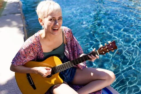 Non-binary person playing acoustic guitar at pool edge, sparkling water Stock Photos
