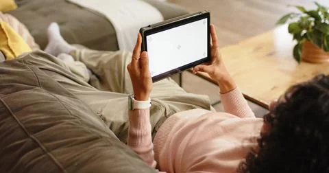 Non-binary person reclining on couch at home tapping blank tablet screen, Stock Photos