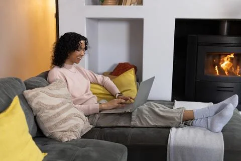 Non-binary person reclining on gray sectional sofa at home typing on laptop Stock Photos