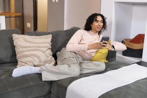 Non-binary person reclining on gray sectional couch at home, holding smartphone Stock Photos