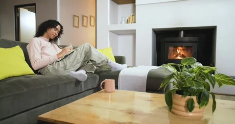 Non-binary person scribbling in notebook on sofa by crackling stove lifting pink Video stock 317589881