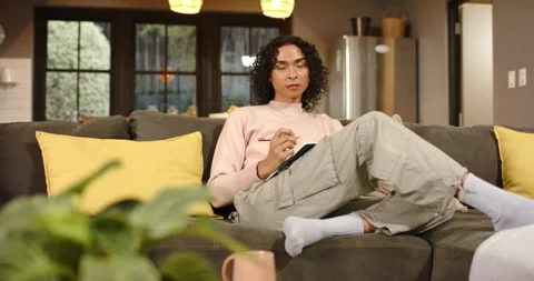 Non-binary person settling on sofa opening notebook, jotting pink pen notes and 動画素材 345123461