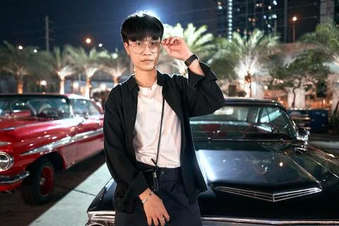 Non-binary person sitting on the bonnet of a luxury car the lot of a night fair Stock Photos