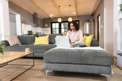 Non-binary person sitting cross-legged on gray ottoman working on laptop in 写真素材