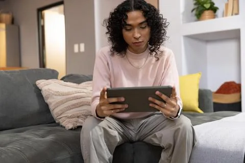 Non-binary person sitting on dark gray sofa in living room holding tablet, Stock Photos