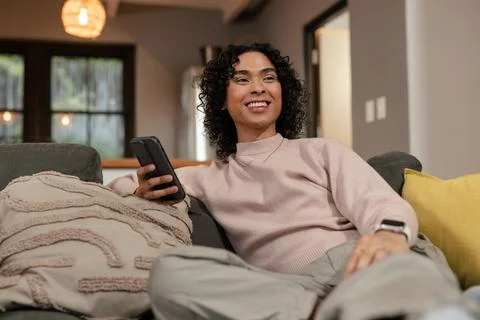 Non-binary person sitting on gray sofa holding smartphone in living room with Stock Photos