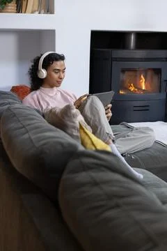 Non-binary person sitting on gray sectional sofa wearing headphones and using Stock Photos