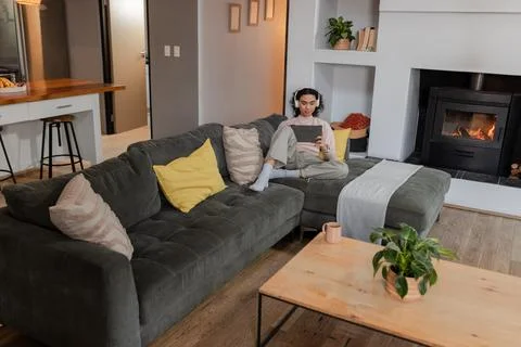 Non-binary person sitting on grey sofa using tablet in living room with Stock Photos