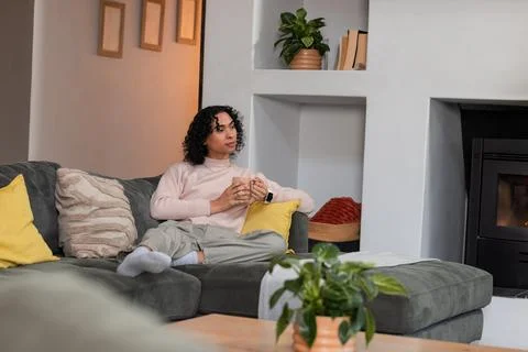 Non-binary person sitting on sofa in living room holding ceramic mug amid Stock Photos