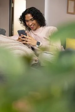 Non-binary person sitting on sofa wearing pink sweater, smiling at smartphone Stock Photos
