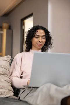 Non-binary person sitting on sofa at home using laptop on lap, wearing pastel Stock Photos