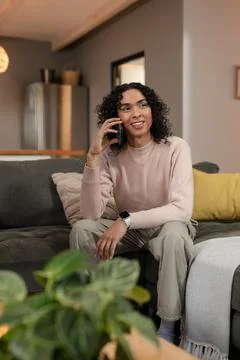 Non-binary person talking on smartphone while sitting on living room sofa with Stock Photos