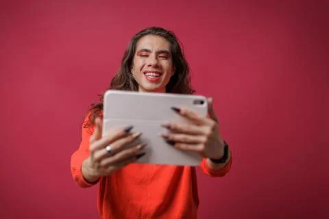 Non-binary person using a tablet on a red background and wearing pride clothi Stock Photos
