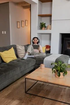 Non-binary person wearing wireless headphones sitting on corner sofa in living Stock Photos
