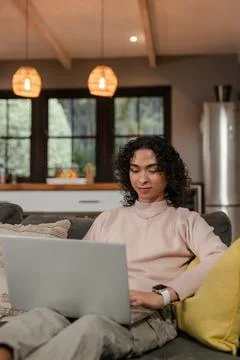 Non-binary person working on laptop on cozy gray sofa in living room with rattan Stock Photos