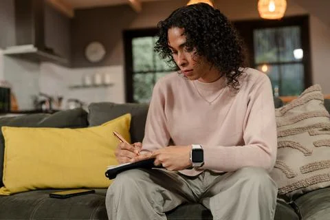 Non-binary person writing in black notebook on gray sofa in home living room Stock Photos