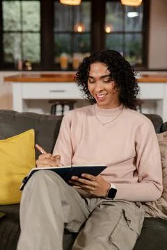 Non-binary person writing in notebook in living area on sofa wearing smartwatch Stock Photos