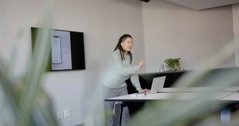 Non-binary presenter tapping laptop, demonstrating smartphone UI on wall display Stock Footage 344673253