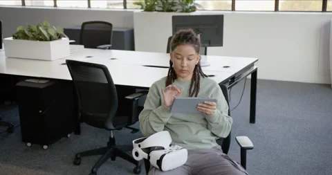 Non-binary sitting in open-plan office holding tablet, tapping to review work, Stock Footage 344693569