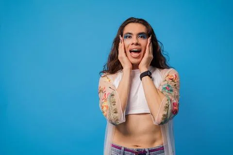 Non-binary stylish person with surprised face and hands on face, blue backg.. Stock Photos