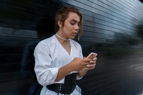 Non-binary using phone in her hands chating leaning on black gate in the street Stock Photos