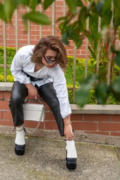 Non-binary vain man fixing his heels shoes with purse in his hands leaning on Stock Photos