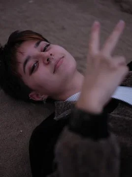 Non-binary woman lying with her head on the sand, making a v-sign with her hand Stock-Fotos