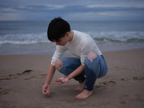 Non-binary woman picking up shells from the sand on the beach Foto stock
