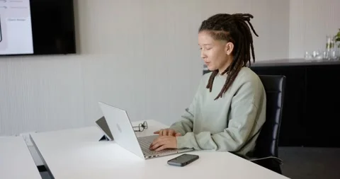 Nonbinary African American typing laptop at long white meeting table, glancing 動画素材 344673246