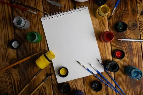 Notebook in the middle with different brushes and gouache colors on a woode.. Stock Photos