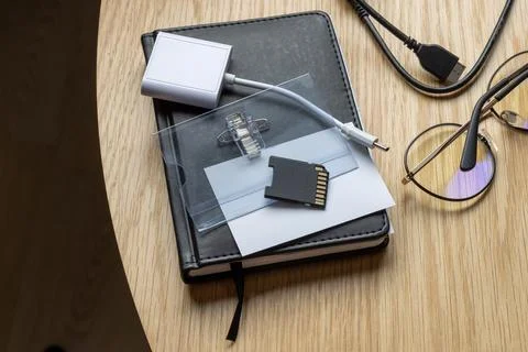 A notebook with a SD card, USB cable, and glasses on a wooden table. 스톡 사진