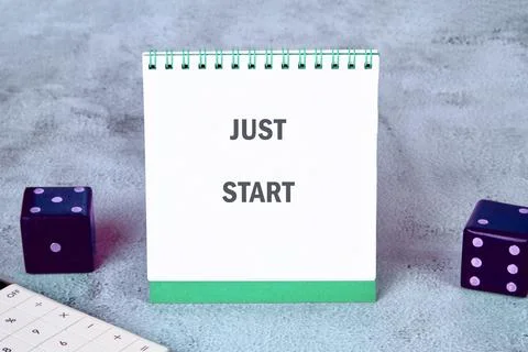 A notepad displays JUST START next to dice and a calculator, on a mottled gray Stock Photos