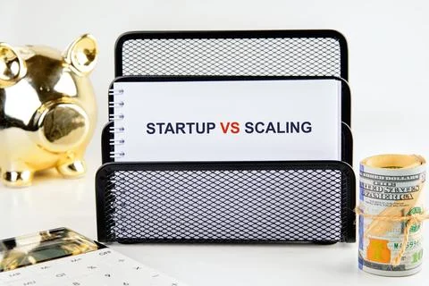 A notepad displays the words startup vs scaling while a calculator, a piggy b Stock Photos