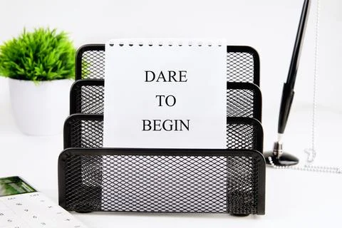 A notepad with the words DARE TO BEGIN sits in a black metal desk organizer. A Stock Photos